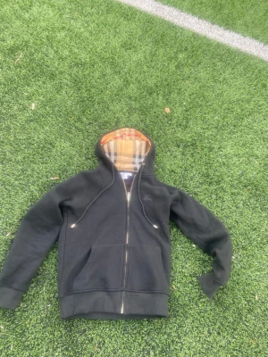 Svart Burberry hoodie med dragkedja - Svart hoodie från Burberry med dragkedja och klassiskt rutigt foder i huvan. Tröjan har fickor framtill, dragsko i huvan och broderad logga på bröstet. Perfekt för en stilren och trendig look. liten i storleken storlek (s)