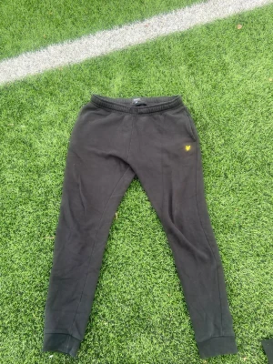 Svarta mjukisbyxor från Lyle & Scott - Svarta mjukisbyxor från Lyle & Scott med gul logga på vänster lår. Byxorna har resår i midjan och muddar vid bensluten. Perfekta för en avslappnad stil och tillverkade i mjukt material för extra komfort.