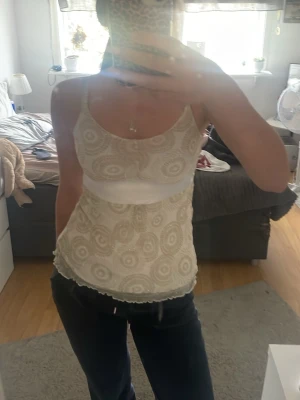 Beige spetsig linnetopp med mönster - Supersöt beige linnetopp med tunna axelband och broderat cirkelmönster. Toppen har en vit satinliknande midjedetalj och vågig spetskant nertill. Perfekt att styla med jeans eller kjol för en trendig look. Storlek 146/152 men väldigt stor i storleken.