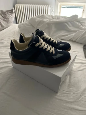 Mörkblå sneakers från Maison Margiela - Snygga mörkblå sneakers från Maison Margiela med vita detaljer och beige sula. Skorna har en klassisk låg siluett, vita snören och är tillverkade i en mix av mocka och läder. Perfekta för dig som gillar stilrena och tidlösa sneakers.