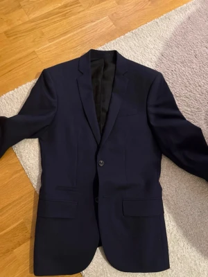 Mörkblå kavaj från Filippa K - Stilren mörkblå kavaj från Filippa K med klassisk passform och två knappar framtill. Kavajen har två fickor med lock och en bröstficka. Fodrad insida för extra komfort. Perfekt för en dressad look.
