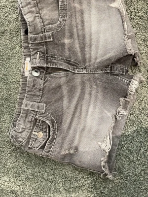 Grå jeansshorts med slitningar - Säljer ett par grå jeansshorts med råa, slitna kanter och dragkedja framtill. Shortsen har klassiska fickor och en cool, tvättad look. Perfekta för dig som gillar en avslappnad och trendig stil.