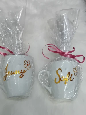 Personliga vita muggar med namn - Två vita porslinsmuggar med guldfärgad text där det står 'Jenny' och 'Sofi', samt en liten blomma bredvid namnen. Muggarna är inslagna i cellofan med rosa band och har en klassisk form med handtag. En mugg kostar 75