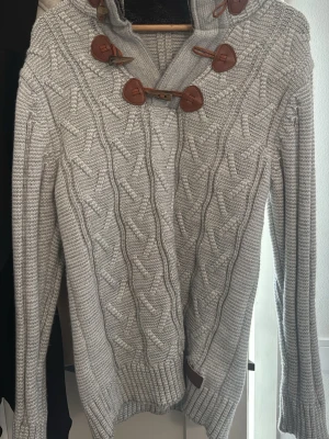 Grå stickad tröja med knappar - Snygg grå stickad tröja med kabelstickat mönster och bruna dekorativa knappar längs halsen. Tröjan har lång ärm och ribbade muddar vid ärmslut och nederkant. Perfekt för kyliga dagar.