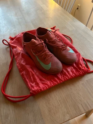 Nike mecurial - Säljer ett par röda Nike fotbollsskor med turkos swoosh. Skorna har snörning och låg profil, perfekt för snabba rörelser på planen. Ovandelen är i syntetmaterial och de kommer med en matchande röd gympåse. Grymma för dig som vill sticka ut på fotbollsplanen! OBS, påse inkluderar!