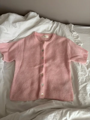 Rosa stickad kofta med kort ärm - Superfin rosa stickad kofta med korta ärmar och rund hals. Koftan har ribbad struktur och knäpps med pärlemorknappar framtill. Perfekt för lager-på-lager och ger en söt och trendig look.