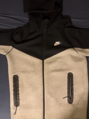 Nike Tech Fleece hoodie svart/grå - Nike Tech Fleece hoodie i svart och ljusgrått med hel dragkedja och två stora fickor med svarta dragkedjor. Klassisk Nike-logga på bröstet och huva för extra stilpoäng. Perfekt för en sportig och avslappnad look.