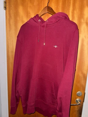 Vinröd hoodie från Gant - Snygg vinröd hoodie från Gant med klassisk huva och dragsko. Liten broderad Gant-logga på bröstet och stor magficka framtill. Tillverkad i mjuk bomullsblandning som är skön att ha på sig. Perfekt för en avslappnad och stilren look.