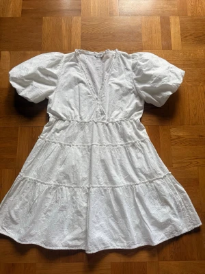 Vit broderad kortklänning med puffärm - Superfin vit kortklänning med broderade blommor och puffärmar. Klänningen har v-ringning, markerad midja och volangdetaljer i kjolen. Perfekt för sommarens alla tillfällen och skolavslutning. Funkar både som M och L har själv M och det sitter jätte fint då också.