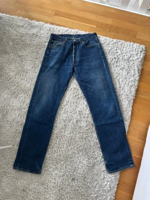 Levi's 501 33/34 raka blå jeans - Klassiska blå jeans från Levi's med rak passform och fem fickor. Jeansen har en snygg tvättad look och detaljer som nitar och läderpatch bak i midjan. Perfekta för en avslappnad och tidlös stil.