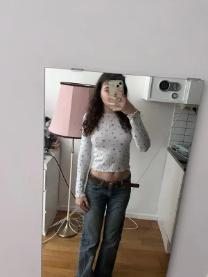 Vit croppad långärmad topp med blommor - Söt vit croppad topp med små blommiga mönster i rött och rosa. Långärmad modell med ribbade kanter och vågig avslutning vid ärmslut och nederkant. Perfekt för dig som gillar en feminin och trendig stil.