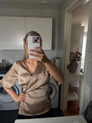 Beige omlottblus  - Snygg beige omlottblus från Bubbelroom i glansigt material. Blusen har knytband i midjan, korta ärmar och djup v-ringning. Perfekt för en trendig och stilren look.