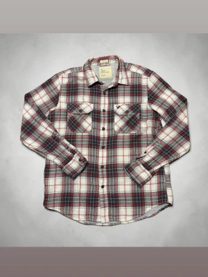 Rutig skjorta från American Eagle - Snygg rutig skjorta från American Eagle Outfitters i rött, blått och vitt. Skjortan har två bröstfickor med knappar och klassisk krage. Tillverkad i mjuk bomull och har lång ärm. Perfekt för en avslappnad och trendig stil.