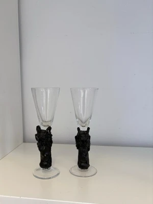 Två glas med svarta djävulshandtag - Säljer två unika glas med svarta handtag formade som djävulshuvuden med horn och skägg. Glasen är genomskinliga upptill och har en cool, detaljerad fot i svart material. Perfekta för dig som gillar annorlunda och kreativa detaljer. En av djävularna är lite sönder