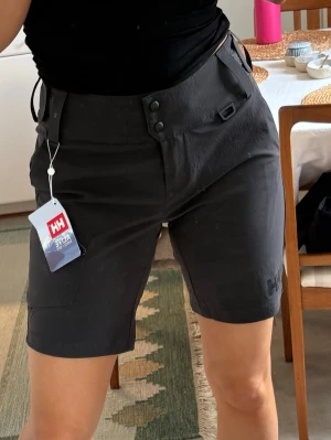 Helly Hansen shorts - Snygga grå shorts från Helly Hansen med hög midja och praktiska fickor. Shortsen har knappstängning framtill och är tillverkade i ett slitstarkt syntetmaterial, perfekta för aktiva dagar. Diskret HH-logga på benet och stilren design.