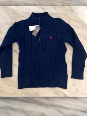 Mörkblå stickad tröja Polo Ralph Lauren - Mörkblå stickad tröja från Polo Ralph Lauren med hög krage och dragkedja. Klassisk kabelstickning och röd broderad logga på bröstet. Tröjan har ribbade muddar och är perfekt för lager-på-lager-stil.