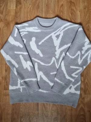 Grå stickad tröja med vitt mönster - Snygg grå stickad tröja med abstrakt vitt mönster över hela plagget. Tröjan har rund halsringning, ribbade muddar och avslut. Perfekt för dig som gillar en modern och avslappnad stil.