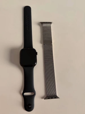 Apple Watch svart med extra armband - Apple Watch med svart silikonarmband och extra silverfärgat mesh-armband i metall. Klockan har en fyrkantig form och digital krona på sidan. Perfekt för dig som vill byta stil snabbt mellan sportigt och stilrent.