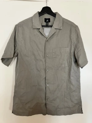 Grön  linneskjorta från H&M - Stilren grön skjorta i linne blandning från H&M med korta ärmar, klassisk krage och bröstficka. Perfekt för en avslappnad look. Skjortan har knäppning framtill och en rak passform. Nypris 299kr. Helt oanvänd.