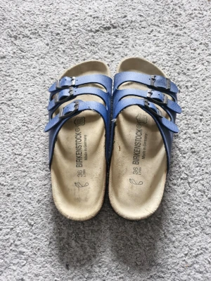 Birkenstock tofflor - Säljer ett par klassiska blå Birkenstock sandaler med tre justerbara remmar och svarta spännen. Sandalerna har en bekväm fotbädd i ljusbeige mocka och är perfekta för en avslappnad stil. Tillverkade i Tyskland. Dem är nästan helt oanvända och är i toppskick. Priset är inte hugget i sten