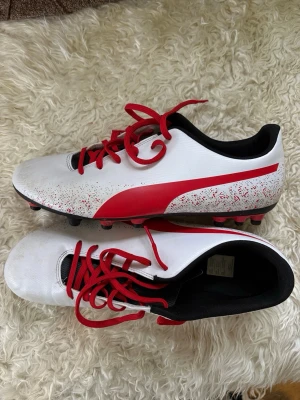 Puma fotbollsskor - Säljer ett par snygga fotbollsskor från Puma i vitt med röda detaljer och röda skosnören. Skorna har klassisk låg profil, svart insida och dobbar för spel på gräs. Materialet är syntetiskt och designen har en stor röd Puma-logga på sidan.