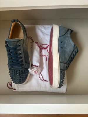 Blåa Christian Louboutin sneakers - Snygga blåa sneakers från Christian Louboutin i mocka med mörkblå snörning och coola nitar på tån. Skorna har den klassiska röda sulan och vita kanter. Perfekta för dig som vill sticka ut med en edgy och exklusiv stil.