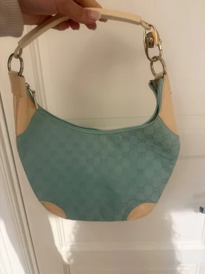 Ljusblå axelväska från Gucci - Säljer en ljusblå axelväska från Gucci med klassiskt GG-mönster i tyget. Väskan har krämvita läderdetaljer på hörn och handtag samt silverfärgade metalldelar. Snygg bågformad design och dragkedja upptill. Perfekt för dig som gillar lyxiga accessoarer. Köpte den i en vintage butik, har inte kommit till användning, Pris går att diskuteras. Skriv om mer bilder 🥰