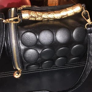 Svart väska med dödskalle detaljer  - 22 cm lång, 17 bredd. En häftig ovanlig svart liten rimlig väska med guldiga dödskalle detaljer på handtaget i jätte fint skick. 2 st dragkedjor så man kan öppna uppifrån och på sidan. 2 små innefickor. Finns axelrem om man vill använda den. 