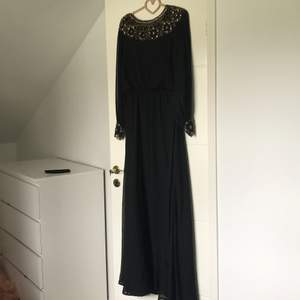 Detta är en lång svart klänning med guldiga detaljer på, lite genomskinligt vid halsen av klänningen men detaljerna täcker för det. 