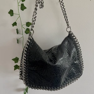 Tiamoväska i svart  - Svart handväska från Tiamo med silvriga detaljer som påminner om Stella McCartney väskorna. Väskan är i fint skick, men spännet baktill har tappat lite färg. Bortsett från detta är det endast normalt slitage. Trots sin storlek rymmer den ganska mycket, på så sätt är den smidig och praktiskt i vardagen. Köparen står för frakten! Detta är en intressekoll och säljer endast vid bra bud!😄