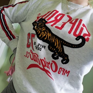 Collegetröja från SuperDry strl. M - Ljusgrå collegetröja med röd, svarta räverer på ärmarna och stort tryck på framsidan av tröjan, helt ny. 