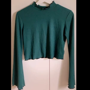 Mörkgrön croppad tröja från NAKD - Jättefin croppad långärmad tröja från NAKD med utsvängda armar och lite ”vågiga” svarta kanter. Materialet är lite ribbat och är jätte mjukt! Det är storlek M men sitter som en S. Köparen står för frakt