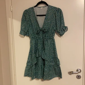 Grön klänning  - Grön klänning med vita blommor och volanger på kjolen och ärmarna samt en rosett. Använd en gång :)