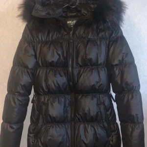 Hollies Jacka med svart päls  - En svart hollies jacka med svart fake päls i storlek 40. Använd ett fåtal gånger så den är i jätte bra skick. Jackan kom med ett svart bälte som jag tyvärr inte hittar men sitter lika fint utan. 