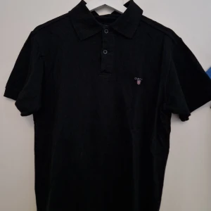 Gant Piké svart - En lite äldre Gant Piké men som fortfarande är i riktigt bra skick. Riktigt hög kvalitet på tyget och som resultat av det så har den klarat sig länge utan slitage. Buda på!