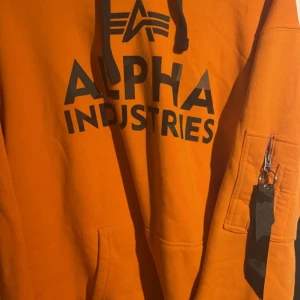 Alpha Industries Hoodie - Ficka på Armen och detaljer i camo färg som tex i luvan och på trycket