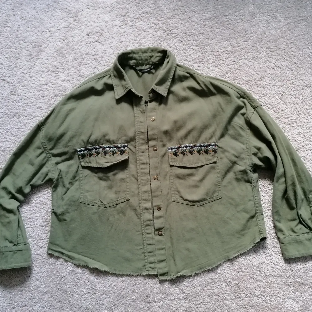 Khaki shirt with frills and embroidery. Oversized fit, can be worn by someone who is between size S - L. Shirt is made of 100% cotton and embroidery 100% polyester. . Jackor.