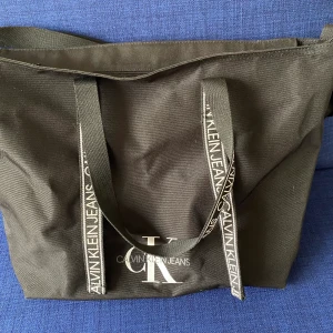 Calvin Klein väska  - Calvin Klein axelremsväska i stark tyg och bra utrymme för träning samt 13 tums laptop. Slitstark en innerficka för plånbok eller mobil. Använt sällan. Snabb affär tack. Köpt på Åhléns 1100kr. Frakt tillkommer 