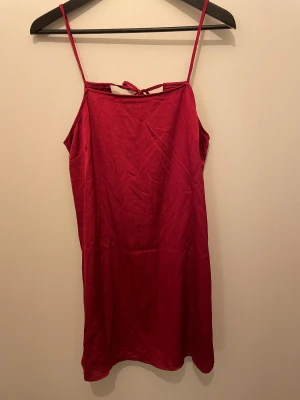 Klänning med öppen rygg - Röd klänning i silkigt material. Öppen rygg med fina knytband. Lite lös i modellen.  Minns ej var den är inköpt ifrån. 