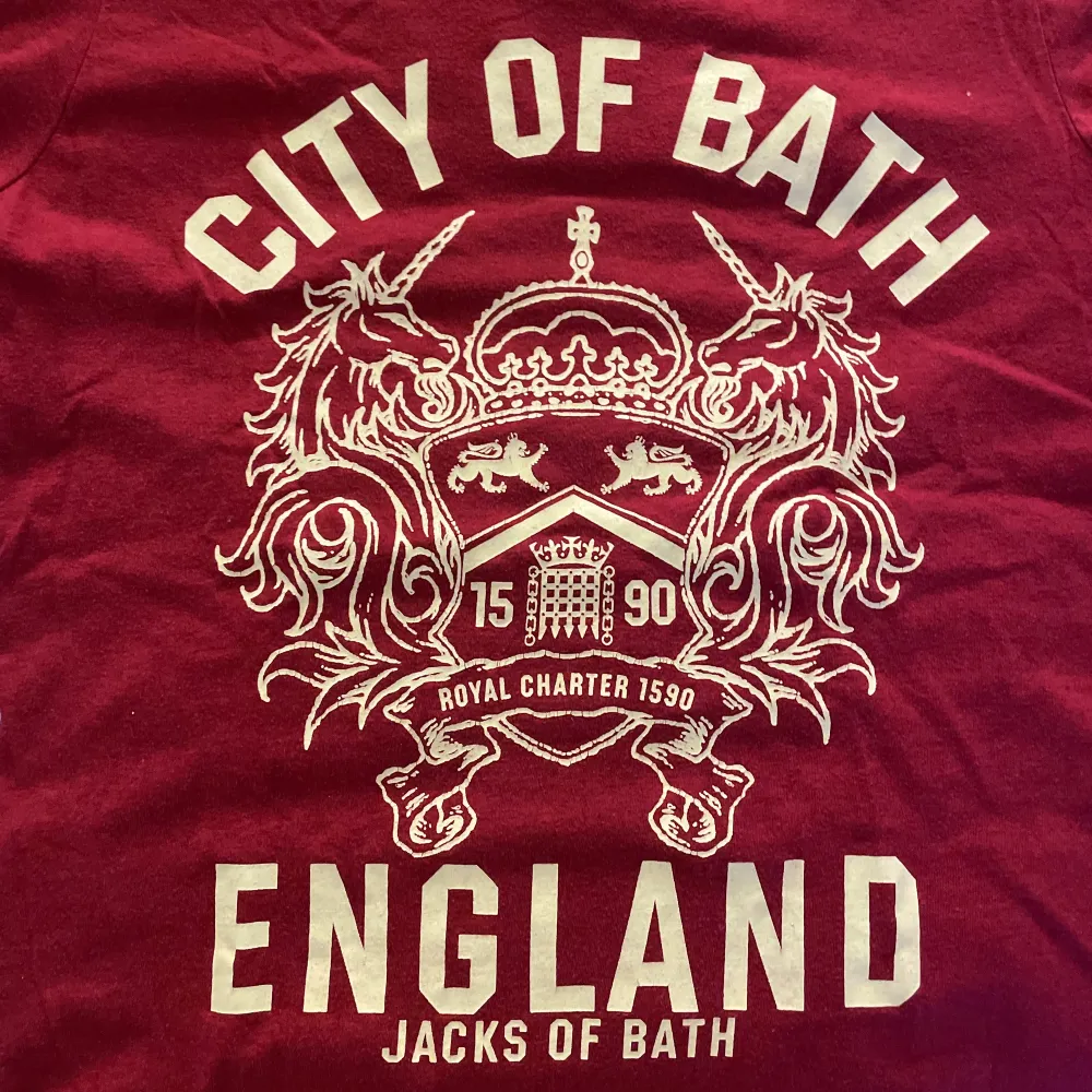 Röd graphic tee köpt på second hand med ett ascoolt gult stort tryck av ett emblem och texten ”City of Bath, England” ❤️⚜️. T-shirts.