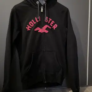 Svart zip up hoodie med rosa Hollister text. Vet ej om den är äkta. Tecken på användning på ena armbågen men inget som syns om man inte kollar nära. Säljer då jag inte tycker om rosa.  Köparen står för frakten.