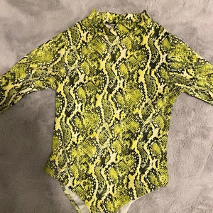 Body - En body i orm mönster, långärmad i färgen neon grön/gul. Du kan knäppa ihop den där nere med knapparna. Hög krage vid halsen.
