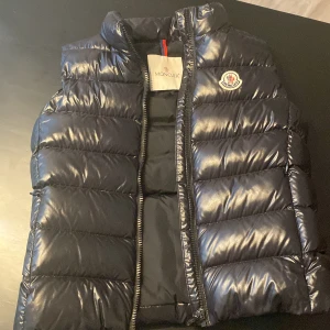 En moncler väst. - En moncler väst i modellen Ghany gilet.Västen är i mycket bra skick jackan har knappast används  och har inga repor eller skador, jackan är köpt från nk i Stockholm, osäker på om kvittot finns kvar men kan garantera äktenbecvis genom Qr-kod, det med kommer också västens etiketter.Pris kan diskuteras❣️