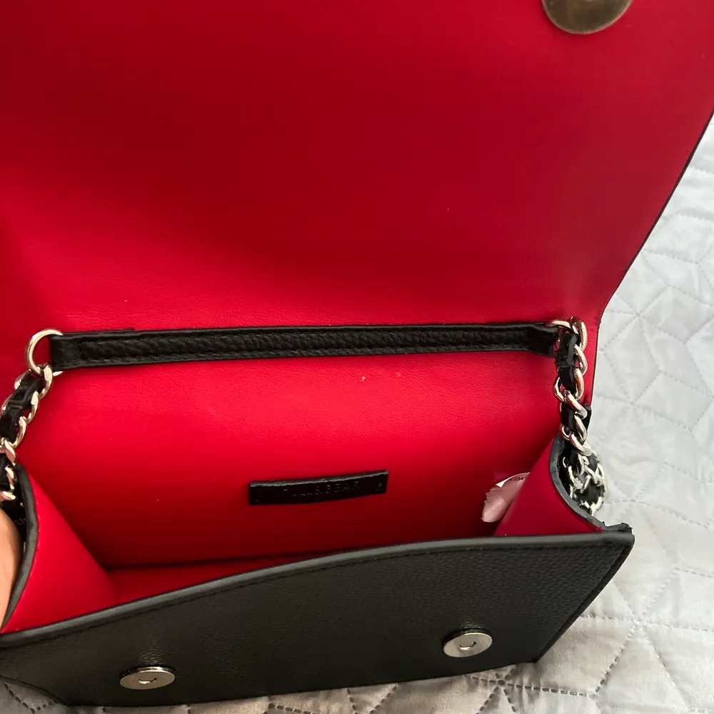 Mindre axelbandsväska i svart med röd insida. Väskan har ett axelband med silvriga detaljer. Använd bara vid 1 tillfälle, väldigt gott skick. . Väskor.