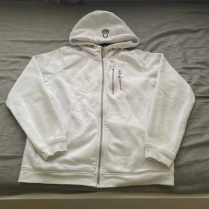 Vit hoodie från Sail Racing - Vit hoodie från Sail Racing med dragkedja hela vägen, broderad logga på bröstet och en extra ficka med dragkedja. Tröjan har huva och långa ärmar, perfekt för chill dagar. Materialet känns mjukt och sportigt. Hör av dig vid frågor mvh Alex 