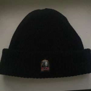 Svart mössa från Parajumpers - Svart ribbstickad mössa från Parajumpers med uppvikt kant och klassisk PJS-logga framtill. Tillverkad i mjukt material som håller dig varm under kyliga dagar. Perfekt accessoar för höst och vinter. Har haft den i 1 månad men den passar inte mig