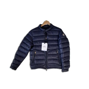 Mörkblå Moncler dunjacka unisex - Snygg mörkblå dunjacka från Moncler med klassisk quiltad design, rund krage och dragkedja framtill. Jackan har två fickor med dragkedja och ikonisk Moncler-logga på ärmen. Perfekt för kyliga dagar och ger en clean, modern look. Det är ett litet hål i en av fickorna, men inget som syns när den används.
