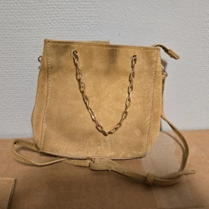 Beige mocka axelväska med kedja - Snygg beige axelväska i mocka med guldfärgad kedja som detalj framtill. Väskan har en enkel, fyrkantig form och justerbar axelrem. Perfekt för dig som gillar stilrena accessoarer med en twist.
