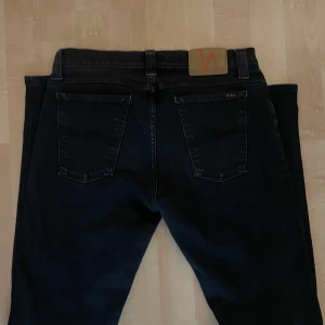Svarta jeans från Nudie Jeans - Svarta jeans från Nudie Jeans med klassisk femficksdesign och snygga detaljer på bakfickorna. Jeansen har en rak passform och är tillverkade i slitstarkt bomullstyg. Perfekta för dig som gillar stilrena och tidlösa jeans.