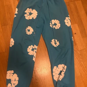 Blå mjukisbyxor från Denim Tears - Blå mjukisbyxor från Denim Tears med vita blommiga tryck över hela byxorna. Byxorna har resår och snörning i midjan för skön passform. Perfekta för en chill och avslappnad stil.